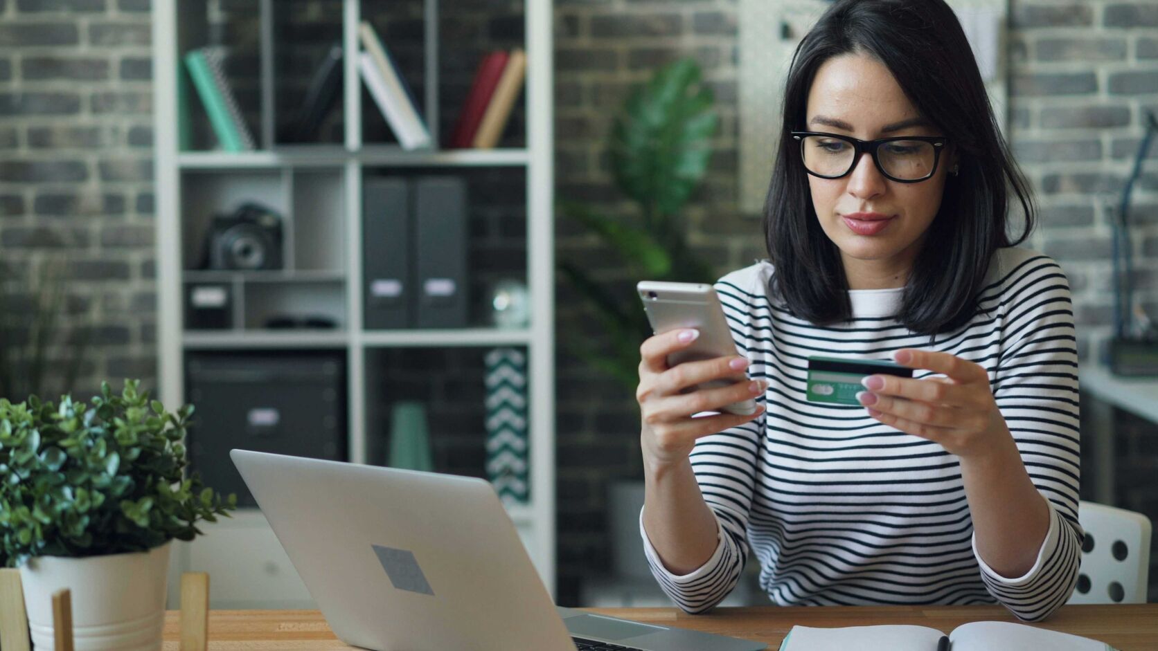 A woman in glasses and a striped top sits at a desk with a laptop, holding a phone in one hand and a payment card in the other.
