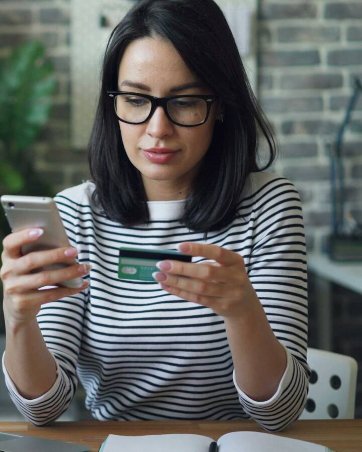 A woman in glasses and a striped top sits at a desk with a laptop, holding a phone in one hand and a payment card in the other.