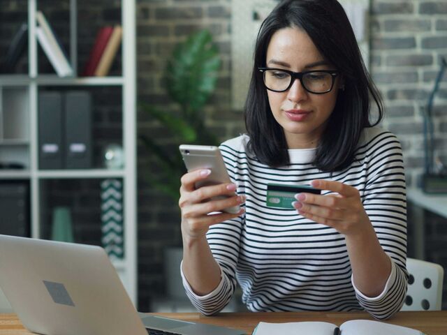 A woman in glasses and a striped top sits at a desk with a laptop, holding a phone in one hand and a payment card in the other.