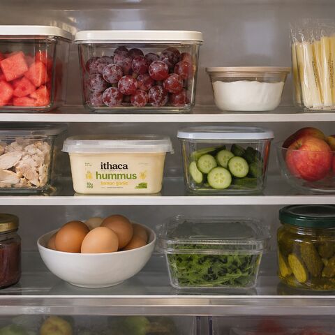 An open refrigerator stocked with first-trimester safe foods — watermelon, grapes, cheese sticks, yogurt, cucumber, apples, hummus, eggs, and pickles