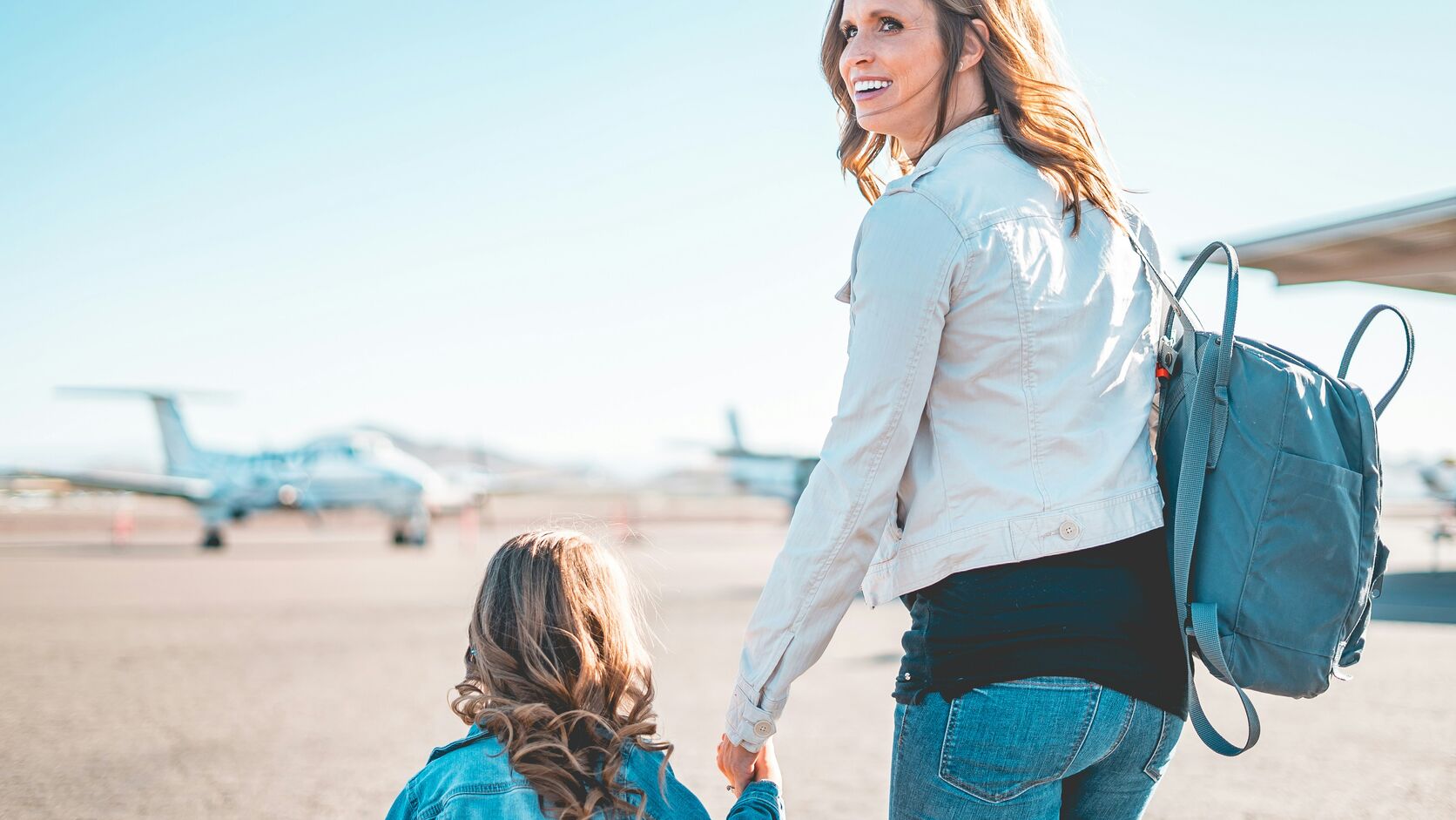 A mother in a light jacket with a blue backpack glances back over her shoulder as she walks hand in hand with her young daughter across an airfield toward a waiting plane.