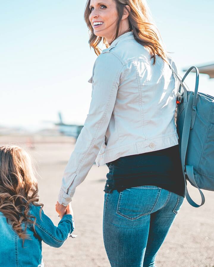 A mother in a light jacket with a blue backpack glances back over her shoulder as she walks hand in hand with her young daughter across an airfield toward a waiting plane.