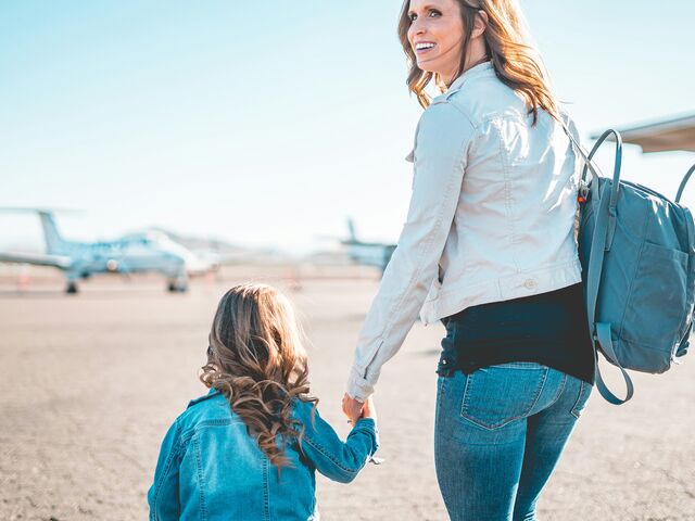 A mother in a light jacket with a blue backpack glances back over her shoulder as she walks hand in hand with her young daughter across an airfield toward a waiting plane.
