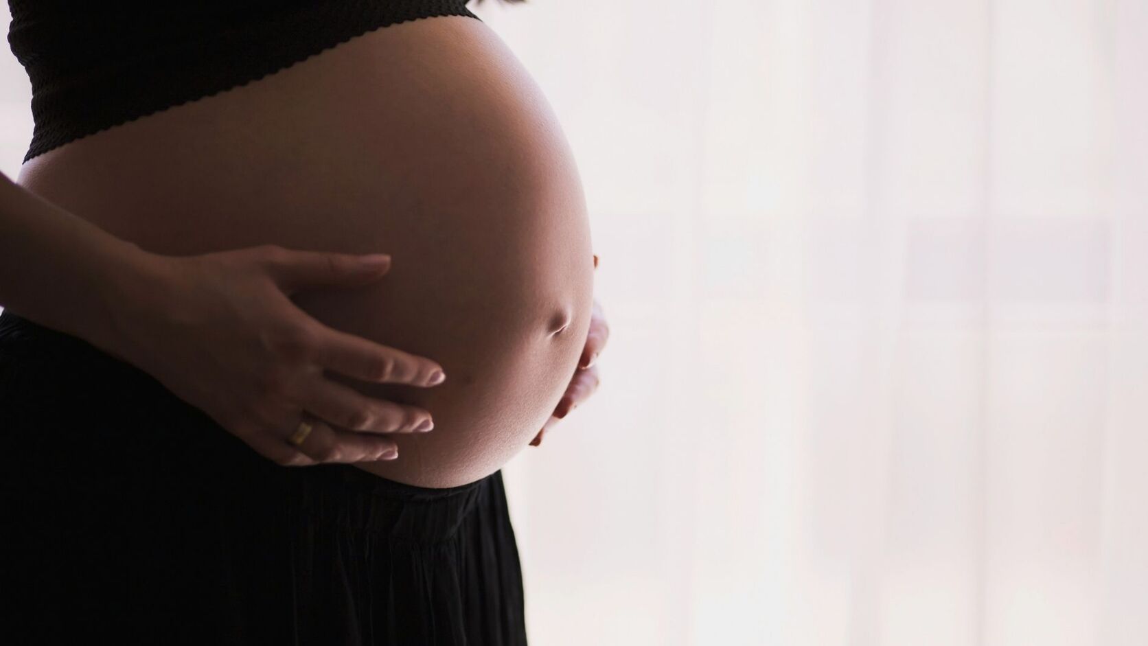 A pregnant woman in black stands in profile by a bright window, both hands cradling her belly.