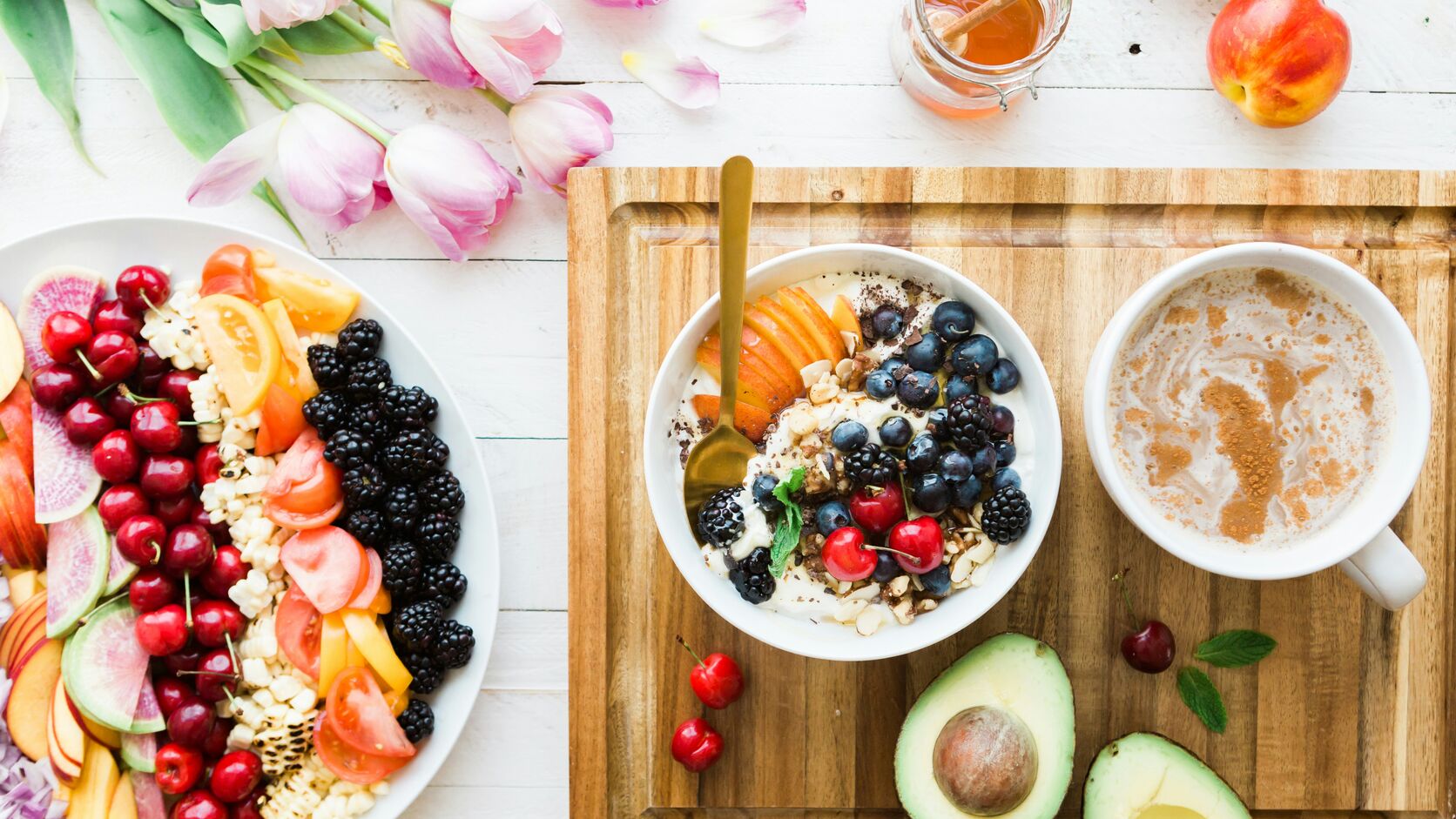 An overhead breakfast spread — a yogurt bowl with berries and granola, a platter of fresh fruit, half an avocado, a warm drink, and tulips on a white table.