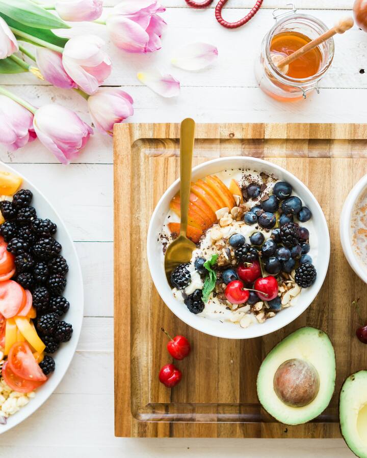 An overhead breakfast spread — a yogurt bowl with berries and granola, fresh fruit, half an avocado, and a warm drink on a white table.