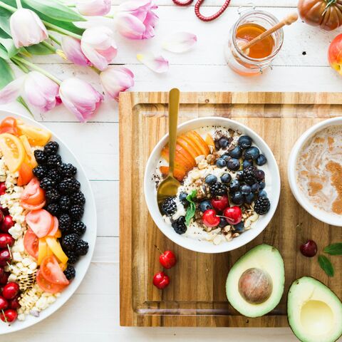 An overhead breakfast spread — a yogurt bowl with berries and granola, a platter of fresh fruit, half an avocado, a warm drink, and tulips on a white table.