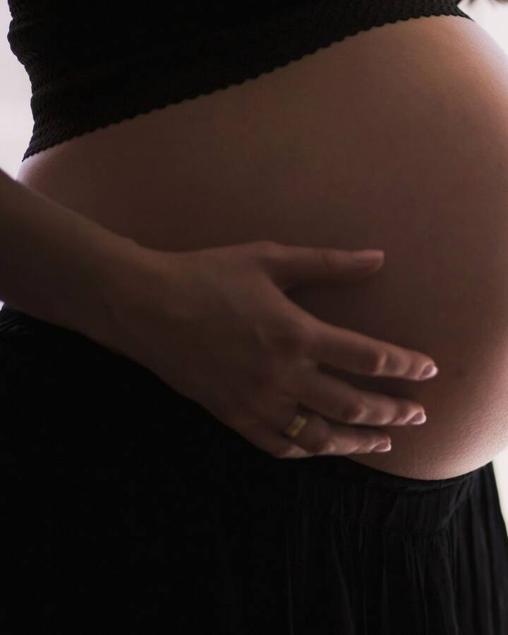 A pregnant woman in black stands in profile by a bright window, both hands cradling her belly.