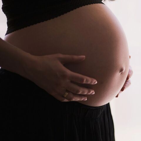 A pregnant woman in black stands in profile by a bright window, both hands cradling her belly.