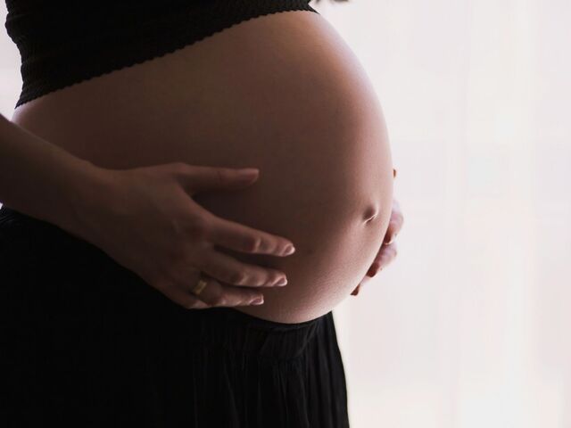 A pregnant woman in black stands in profile by a bright window, both hands cradling her belly.