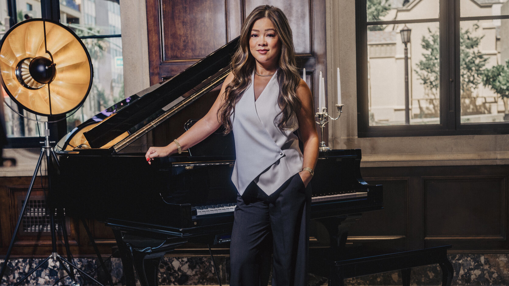 Geraldine Pingul, in a sleeveless pale grey wrap top and wide black trousers, stands with one hand resting on a black grand piano and the other in her pocket, beside a gold studio lamp and tall windows in a wood-panelled room.