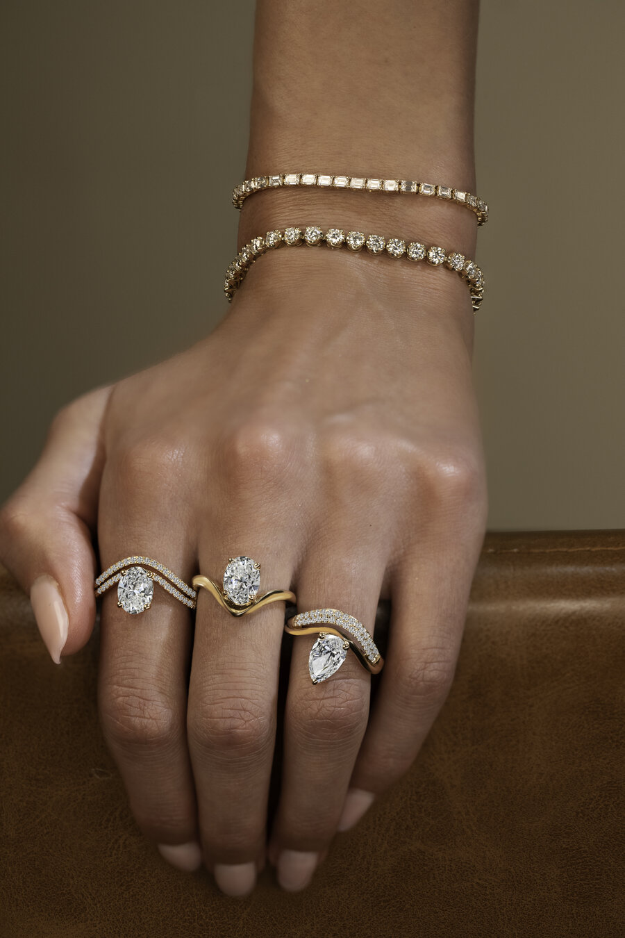 A hand rests on a tan leather chair back, wearing two diamond tennis bracelets in yellow gold and three diamond engagement rings, two ovals and a pear, each paired with a curved diamond or gold band.
