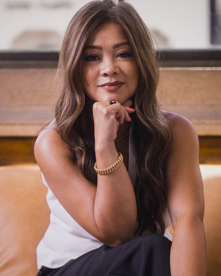 Geraldine Pingul, in a white sleeveless top with long, wavy highlighted hair, rests her chin on her fist and smiles at the camera, a gold chain bracelet on her wrist and a black-and-diamond ring on her finger.