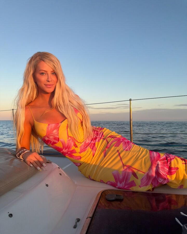 Gigi Gorgeous reclines on the bow of a sailboat at sunset in a yellow and pink floral maxi dress, the ocean behind her.