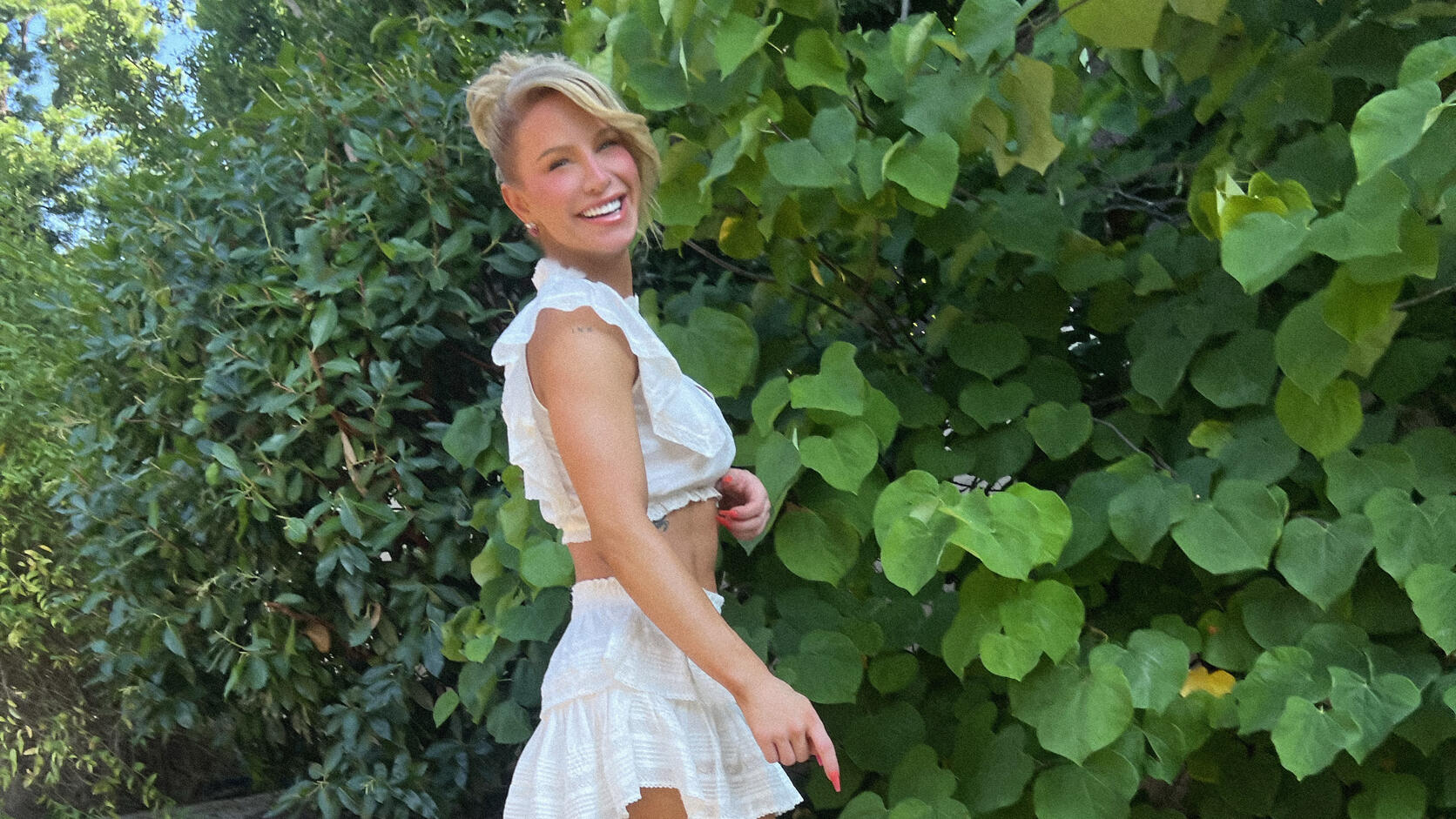 Gigi Gorgeous walks along a garden path in a white ruffled crop top and skirt, laughing over her shoulder in front of a wall of green leaves.