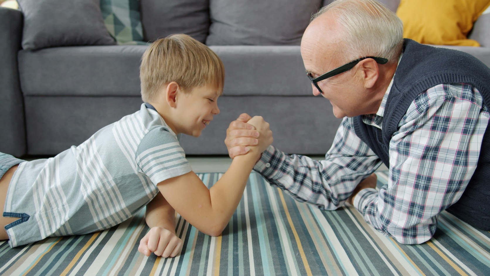 A grandfather in glasses and a plaid shirt arm-wrestles his grinning grandson as they lie on their stomachs on a striped rug in front of a grey sofa.