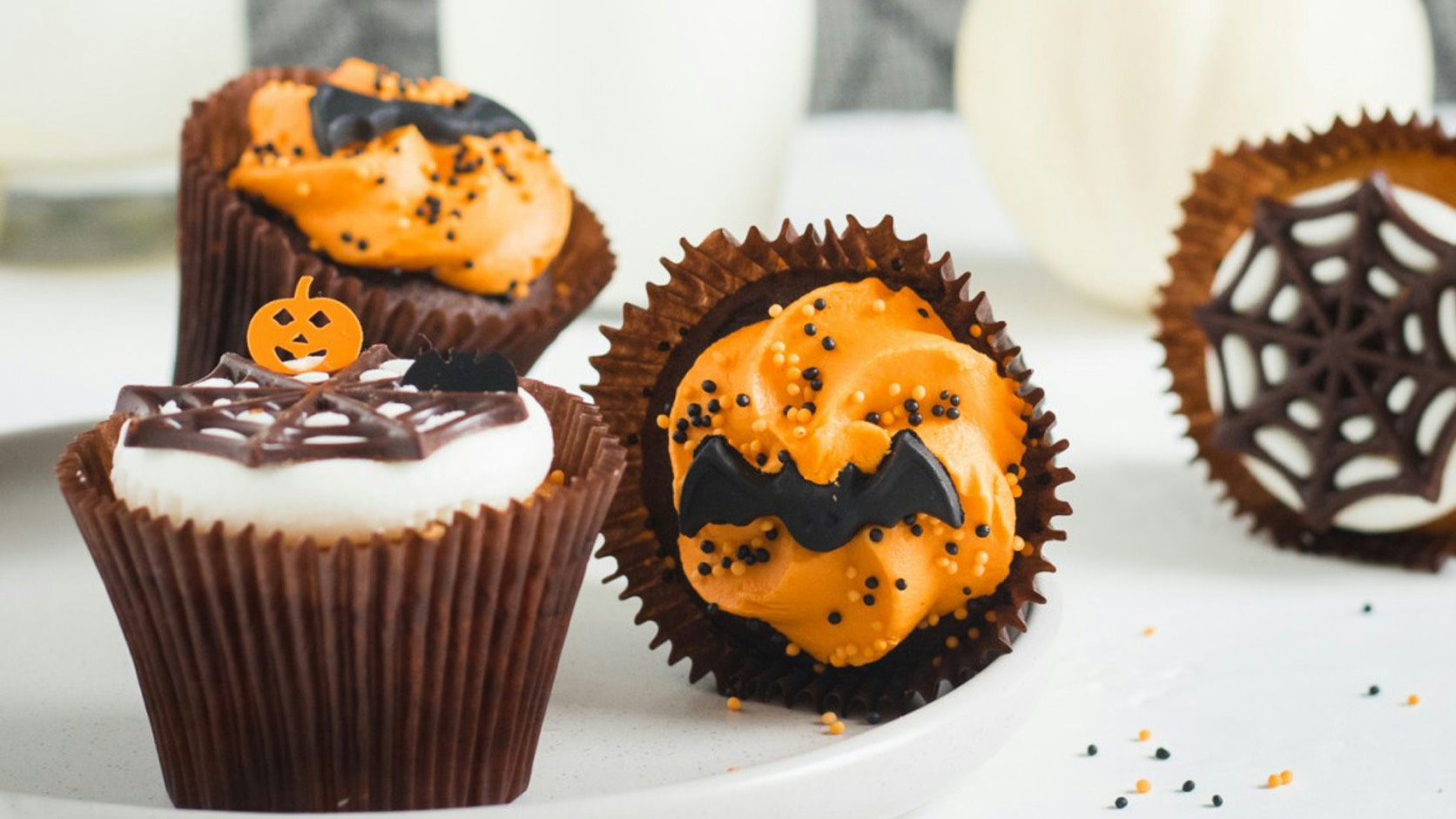 Halloween cupcakes on a white plate, topped with swirls of orange frosting, black bat toppers, orange and black sprinkles and chocolate spiderweb decorations, with glasses of milk and a small white pumpkin behind them.