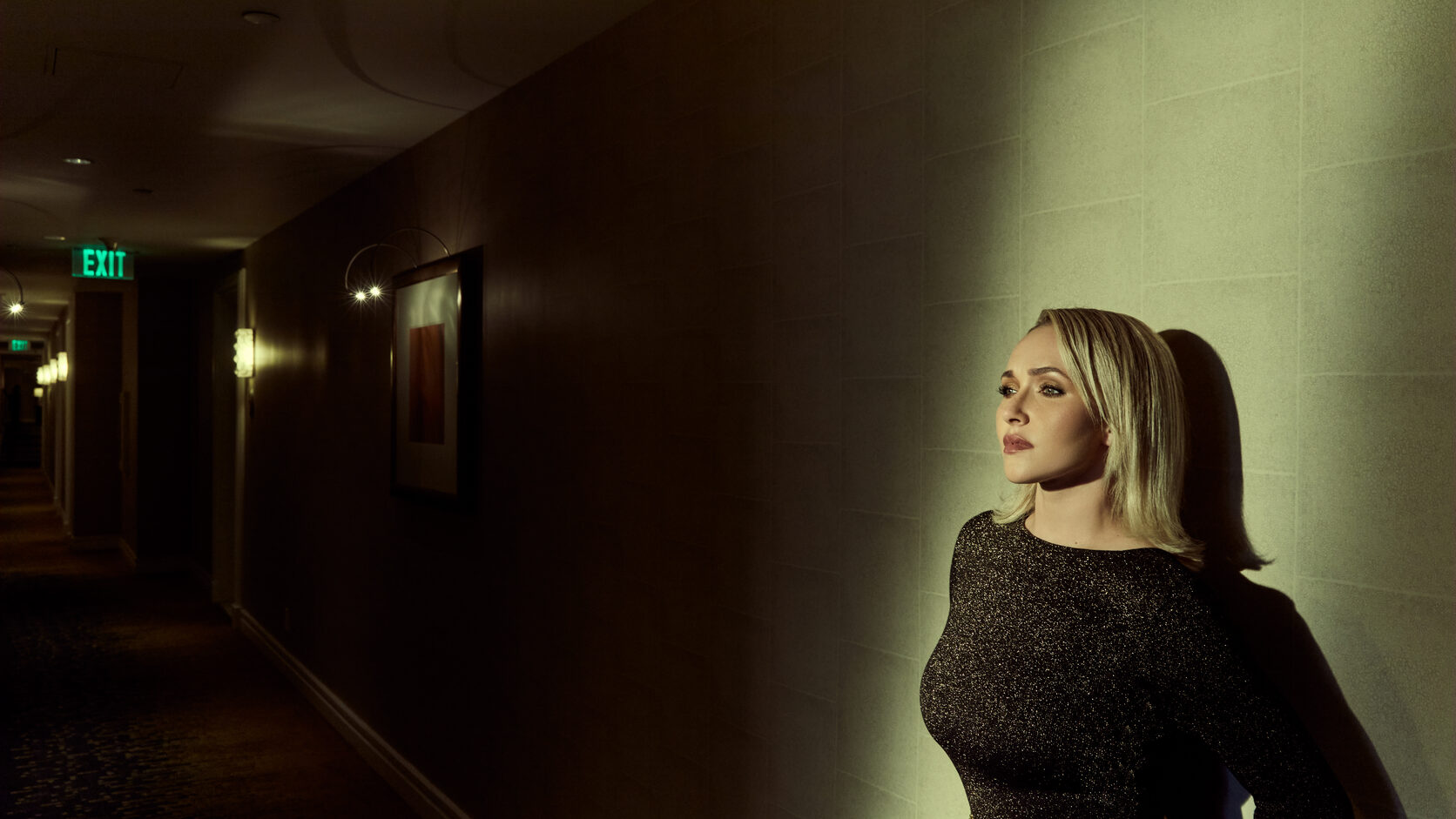 Hayden Panettiere stands against a softly lit wall in a dim hotel corridor, wearing a black long-sleeved dress with a gold medallion belt, gazing off to the side as a green EXIT sign glows in the shadows behind her.