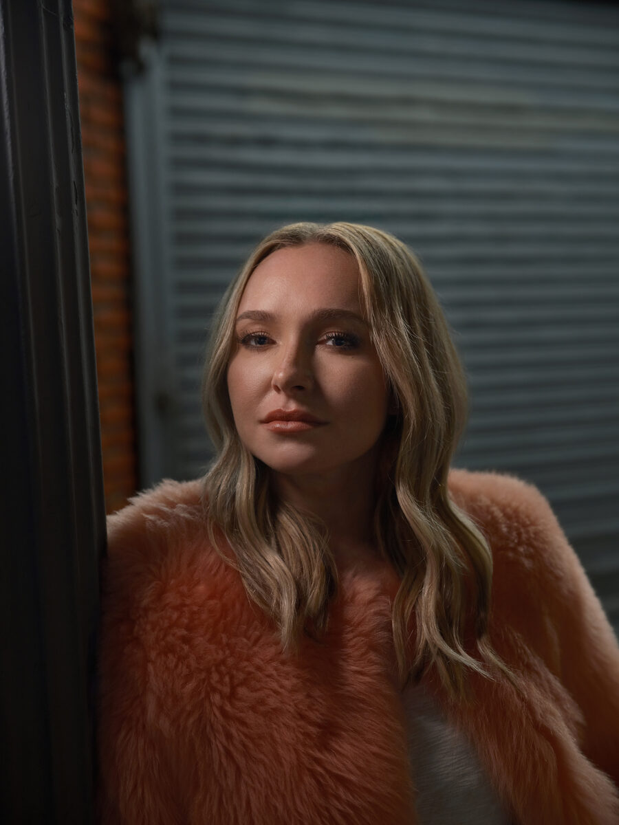 Hayden Panettiere in a peach fur coat leans against a doorway at night, her hair down, looking directly at the camera with a calm, steady expression.