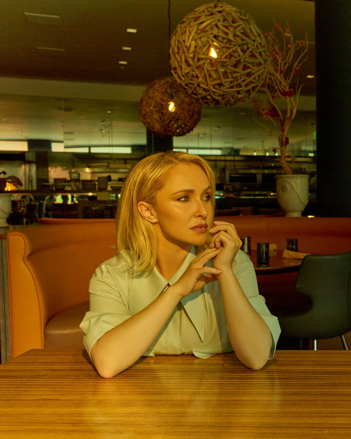 Hayden Panettiere seated alone at a restaurant table in a pale mint shirt, her chin resting on her folded hands as she gazes thoughtfully into the distance beneath warm woven pendant lights.