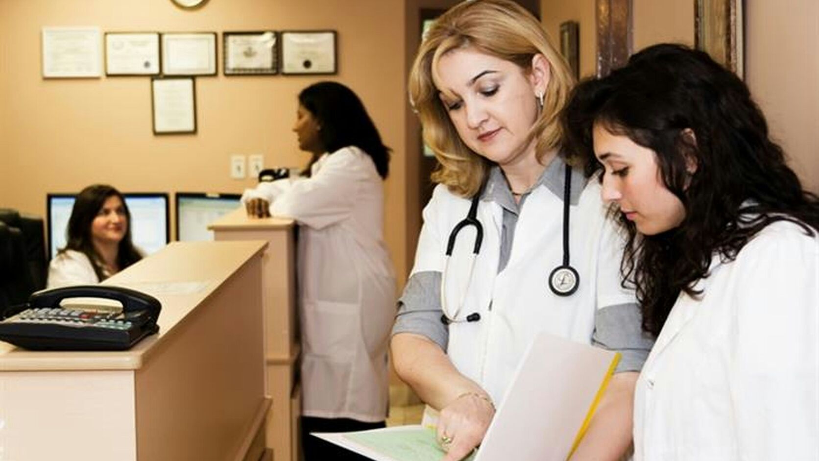 A doctor in a white coat with a stethoscope reviews a chart alongside a younger colleague at a clinic reception desk.