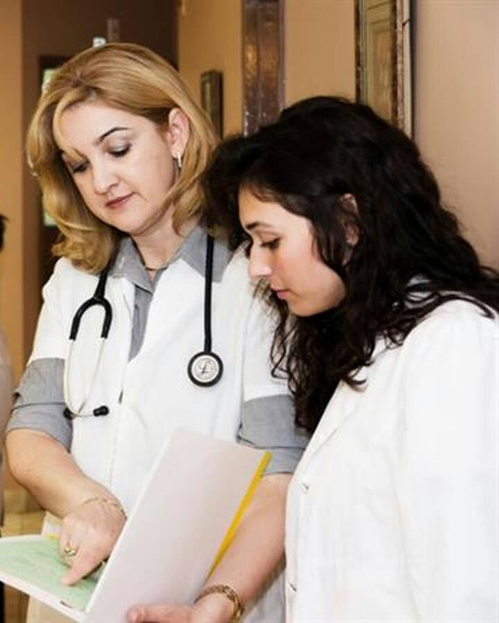 A doctor in a white coat with a stethoscope reviews a chart alongside a younger colleague at a clinic reception desk.