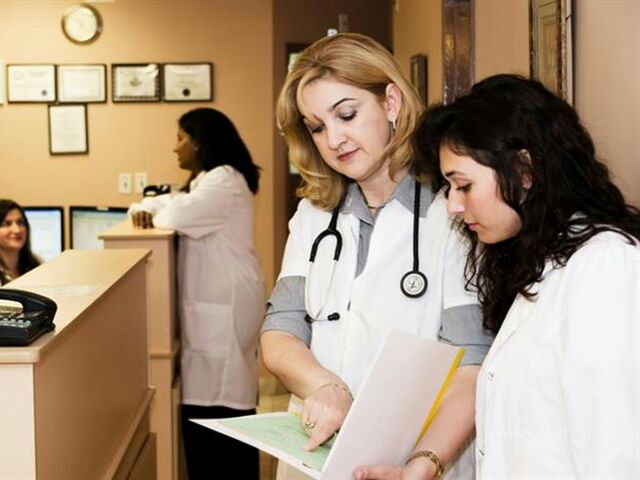 A doctor in a white coat with a stethoscope reviews a chart alongside a younger colleague at a clinic reception desk.