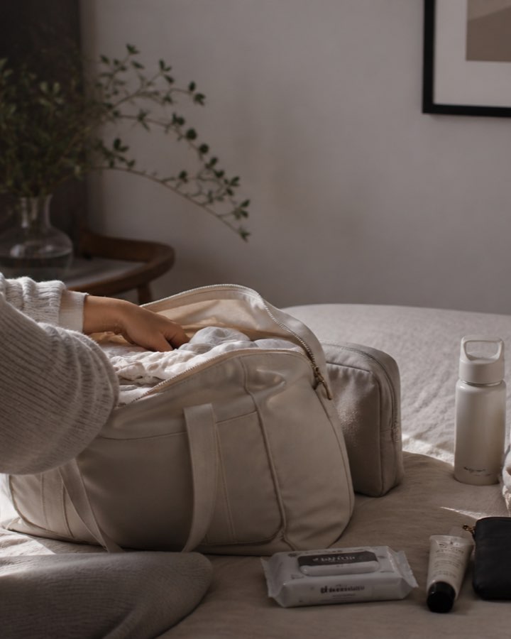 A woman packing a soft cream weekender bag on a bed, with a water bottle, folded linens, and a toiletry pouch laid out beside it in a calm neutral bedroom