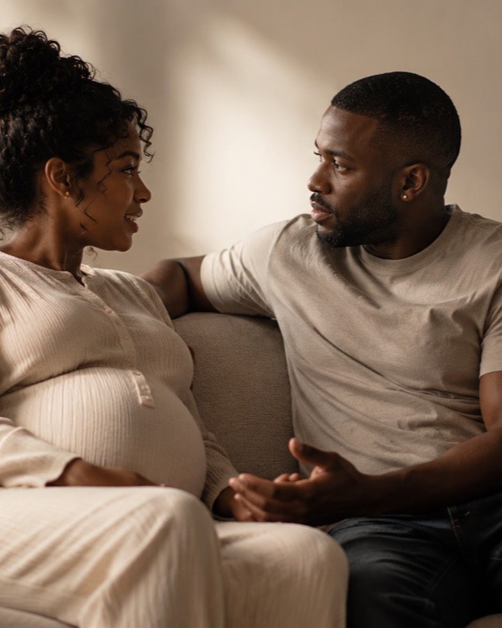 A pregnant woman and her partner sit close together on a cream sofa in soft light, turned toward each other mid-conversation.