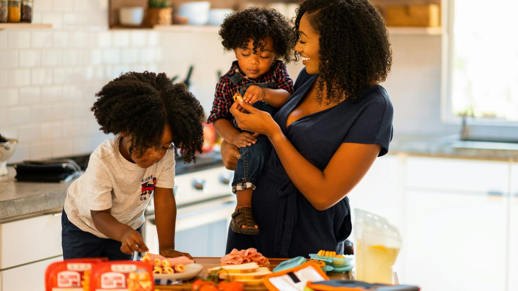 A smiling mother holds her toddler on one hip while her older child helps assemble food at a sunny kitchen counter laid with deli meats, vegetables, and fruit.