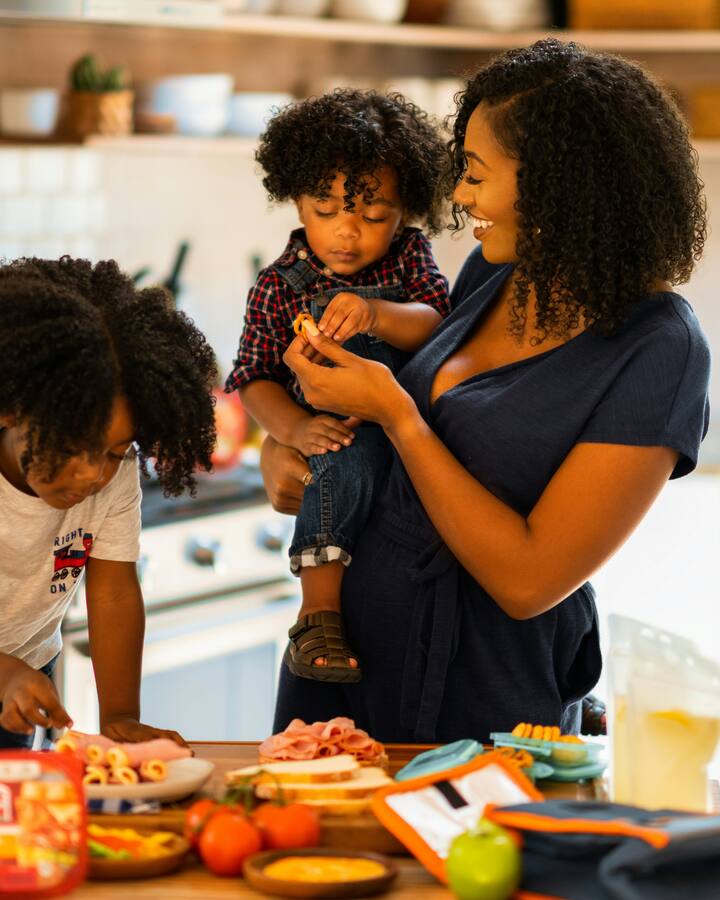 A smiling mother holds her toddler while her older child helps assemble food at a kitchen counter.