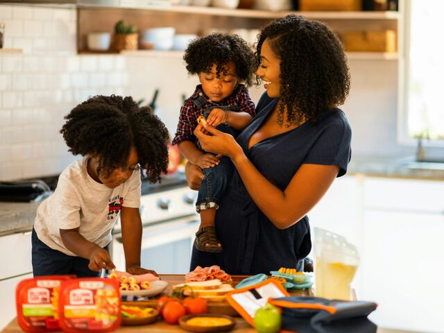 A smiling mother holds her toddler while her older child helps assemble food at a kitchen counter.