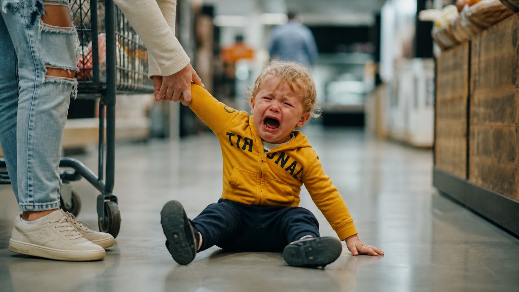 A blonde toddler in a mustard hoodie sits crying on a supermarket floor, holding their mother's hand beside a shopping cart in the grocery aisle.