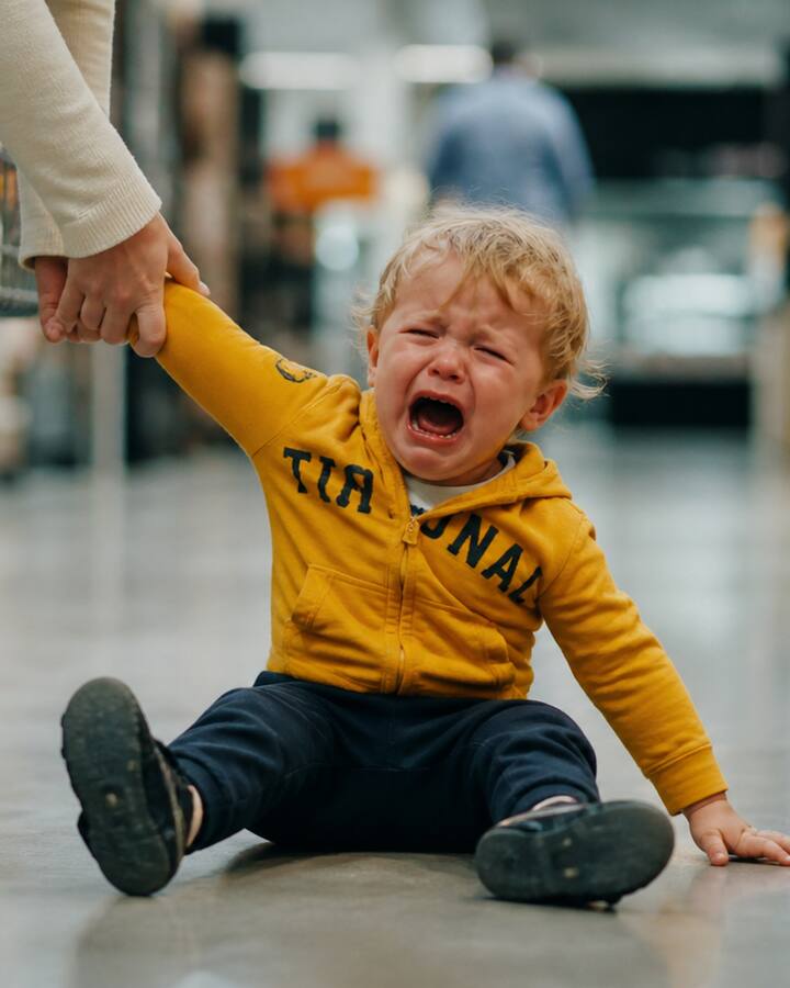 A blonde toddler in a mustard hoodie sits crying on a supermarket floor, holding their mother's hand beside a shopping cart.