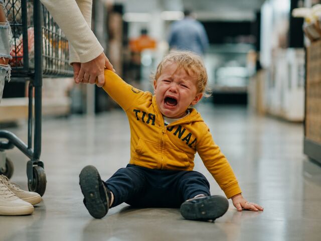 A blonde toddler in a mustard hoodie sits crying on a supermarket floor, holding their mother's hand.