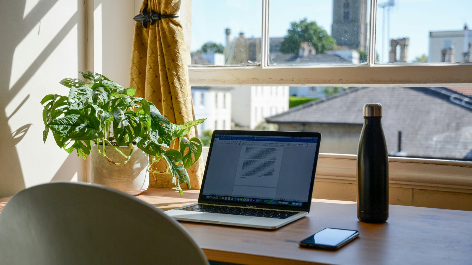 A laptop open on a sunlit wooden desk beside a leafy plant, a water bottle, and a phone, looking out a window over rooftops.