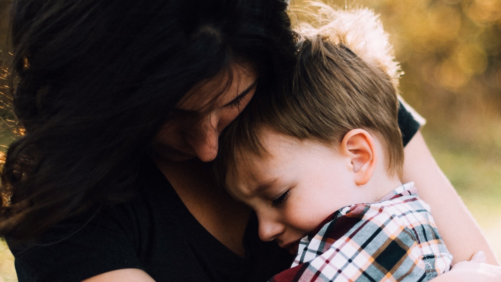 A mother with long dark hair holds her young son close in her lap outdoors, her head bowed against his as he leans into her chest with his eyes closed.