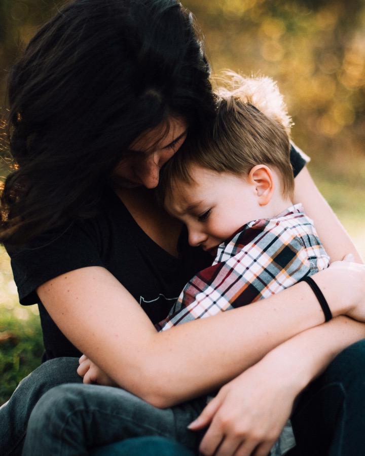 A mother with long dark hair holds her young son close in her lap outdoors, her head bowed against his as he leans into her chest with his eyes closed.