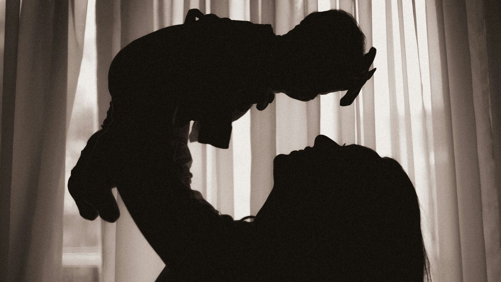 A mother in silhouette lifts her baby high above her face, both backlit by sheer curtains glowing in a bright window.