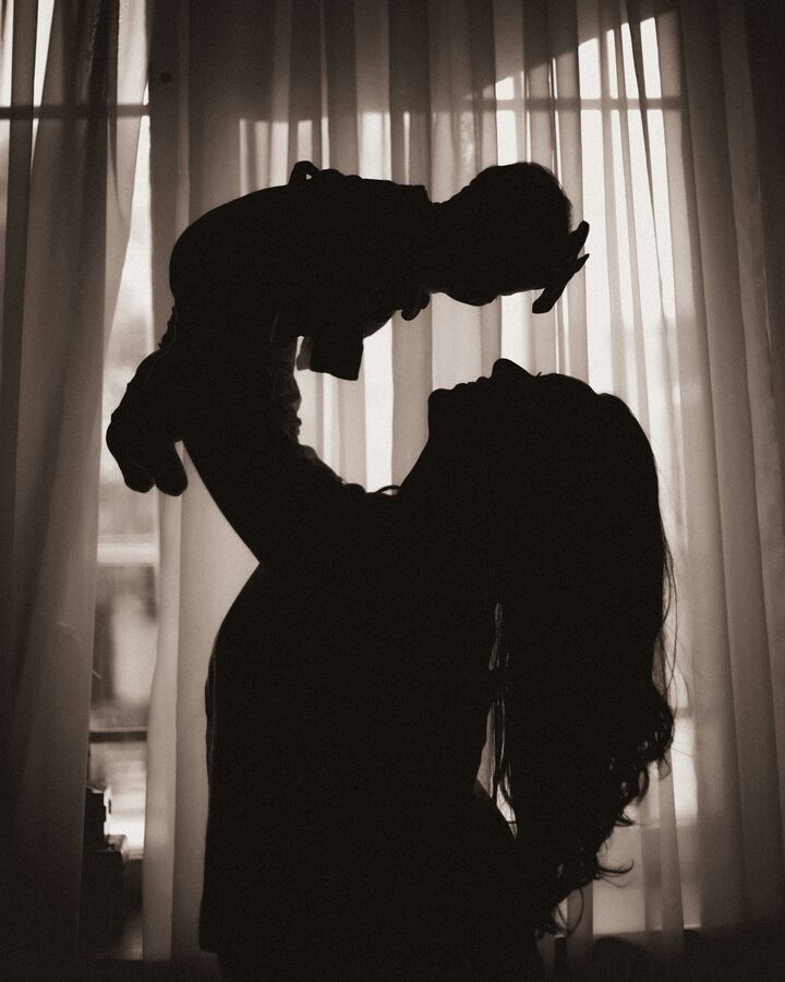 A mother in silhouette lifts her baby high above her face, both backlit by sheer curtains glowing in a bright window.