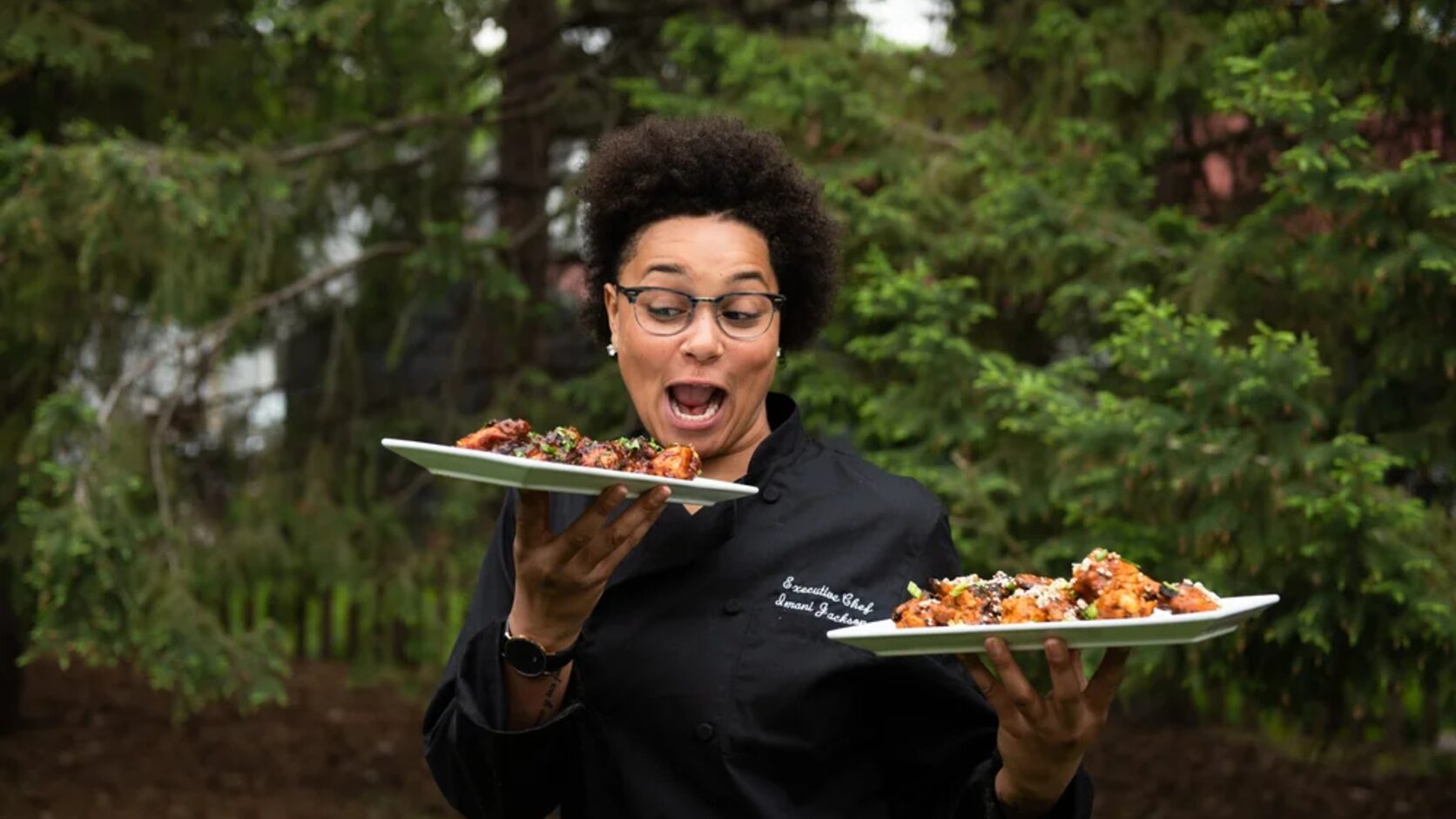 Chef Imani Jackson stands outdoors in a black chef's coat, delightedly eyeing two platters of saucy, garnished food she holds in each hand, green trees behind her.