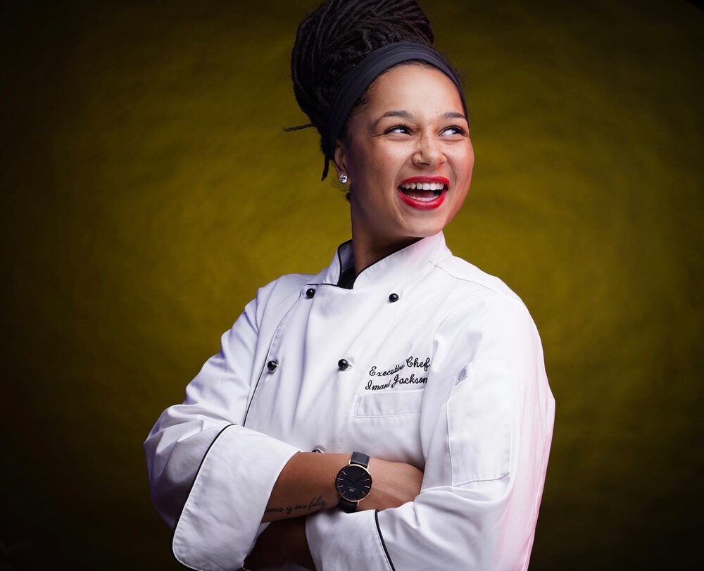 Chef Imani Jackson laughs and looks off to the side in a white chef's coat embroidered 'Executive Chef Imani Jackson,' against a warm olive backdrop.