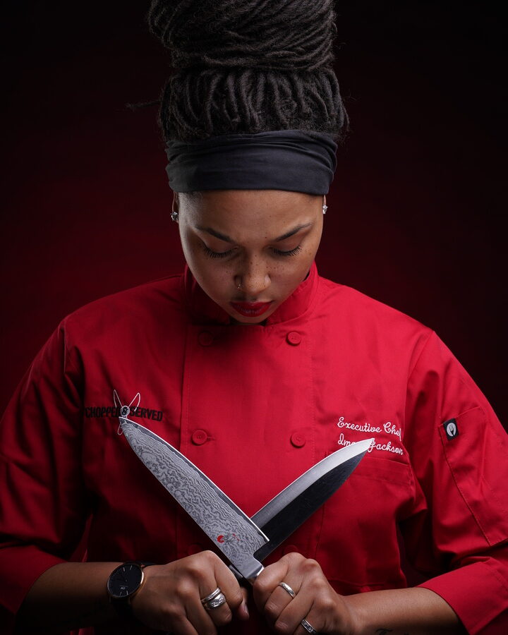 Chef Imani Jackson in a red chef's coat holds two crossed chef's knives, looking down, against a deep red backdrop.