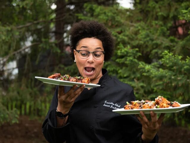 Chef Imani Jackson stands outdoors in a black chef's coat, delightedly eyeing two platters of saucy, garnished food she holds in each hand, green trees behind her.
