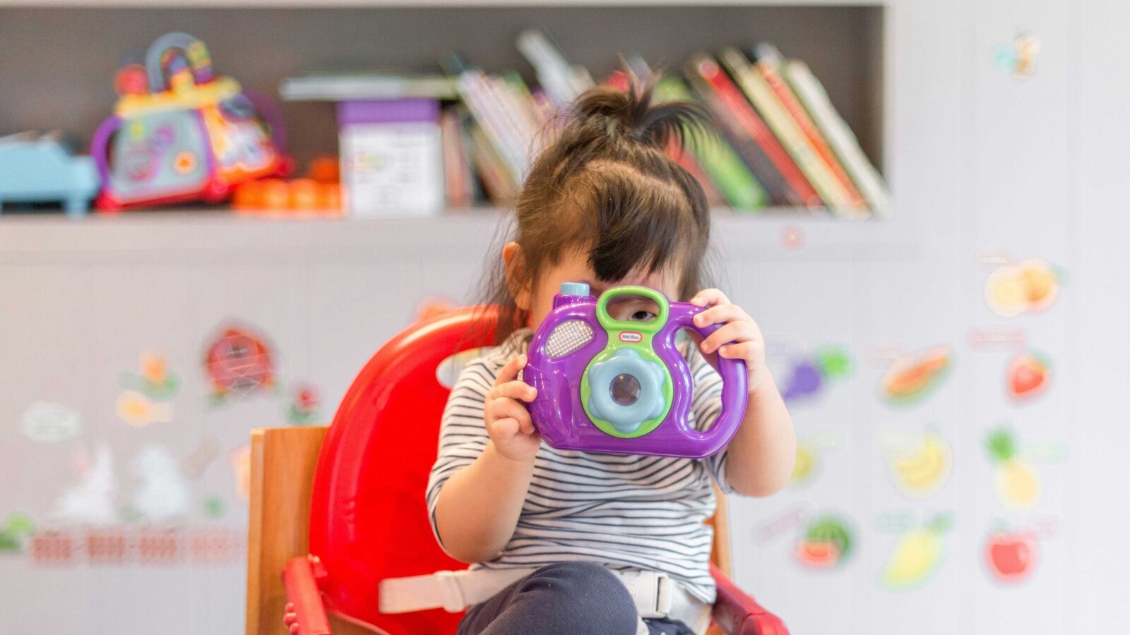 A toddler in a striped shirt sits in a red high chair holding a purple toy camera up to their face, in a colourful playroom.