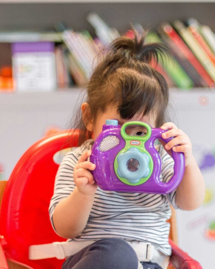 A toddler in a striped shirt sits in a red high chair holding a purple toy camera up to their face, in a colourful playroom.