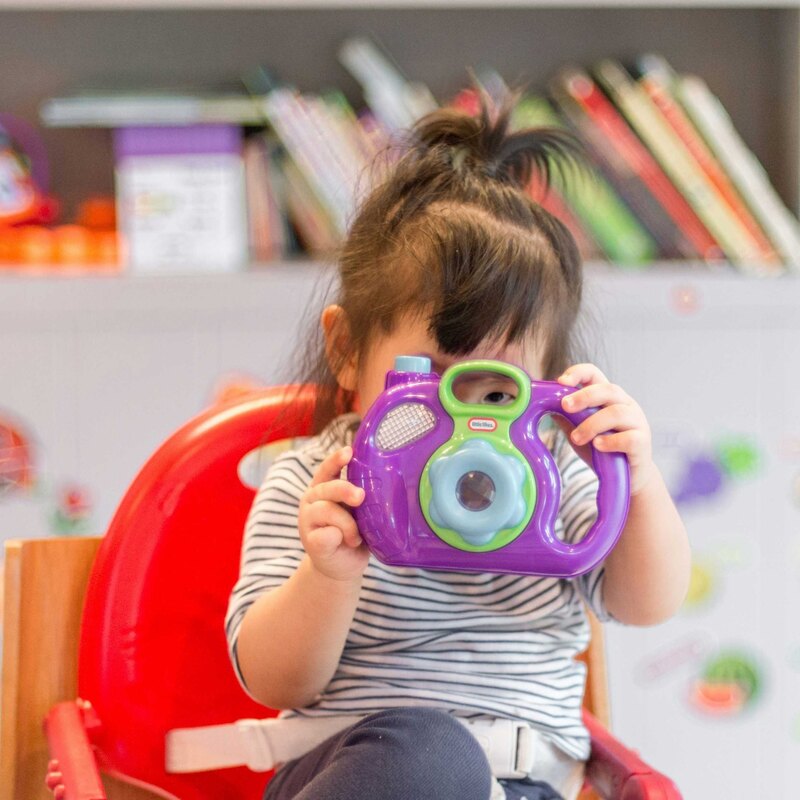A toddler in a striped shirt sits in a red high chair holding a purple toy camera up to their face, in a colourful playroom.
