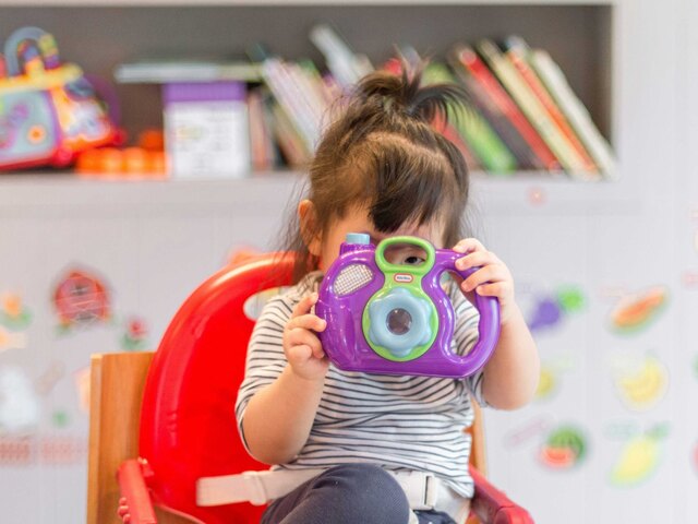 A toddler in a striped shirt sits in a red high chair holding a purple toy camera up to their face.