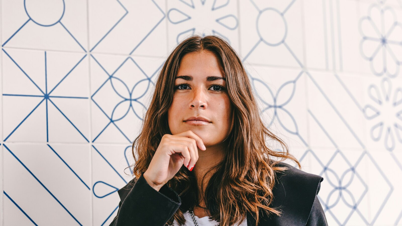 A woman in a dark blazer rests her chin on her hand, gazing thoughtfully, against a white wall patterned with blue tilework.