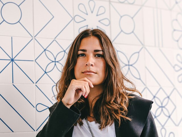 A woman in a dark blazer rests her chin on her hand, gazing thoughtfully, against a white wall patterned with blue tilework.